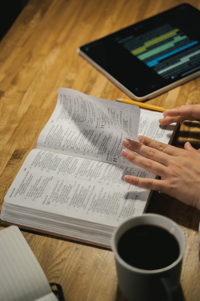 Close-up of hands turning Bible pages with a coffee cup and tablet on the wooden table.